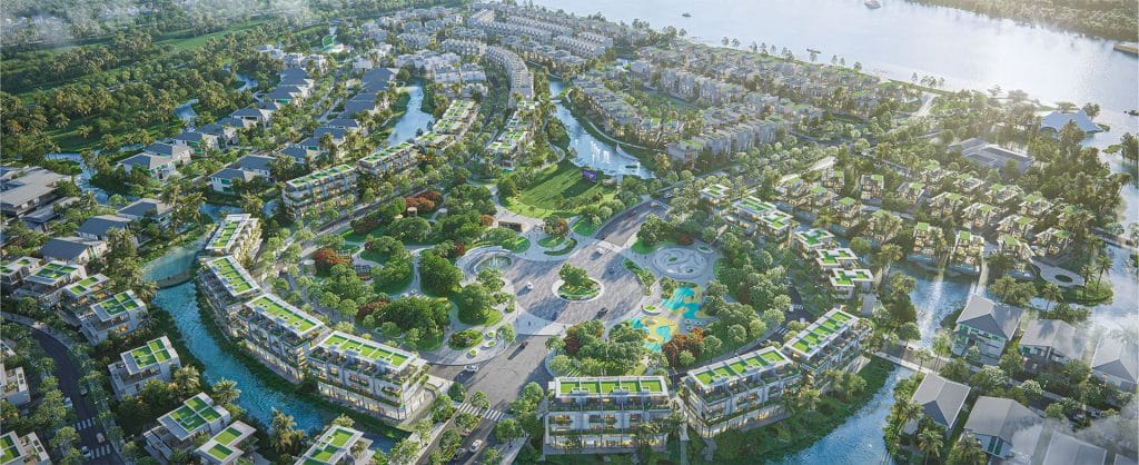 Hình ảnh toàn cảnh dự án Alluvia City từ trên cao, thể hiện một khu đô thị hiện đại với các tòa nhà mái xanh, đường phố uốn lượn, công viên trung tâm và dòng sông chảy qua, bao quanh bởi cây cối và không gian xanh mát. Một hồ nước lớn nằm ở phía bên phải, phản chiếu ánh sáng mặt trời, tạo nên khung cảnh hài hòa giữa thiên nhiên và kiến trúc.