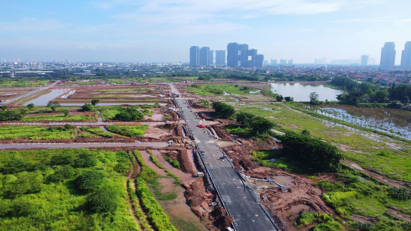 Ảnh chụp từ trên cao toàn cảnh trục đường chính của dự án Alluvia City, với mặt đường đã được trải nhựa, hai bên có các ô đất phân khu đang thi công dở dang, nền đất ẩm và nhiều vũng nước còn đọng; phía xa là khu dân cư và các tòa nhà cao tầng hiện hữu.