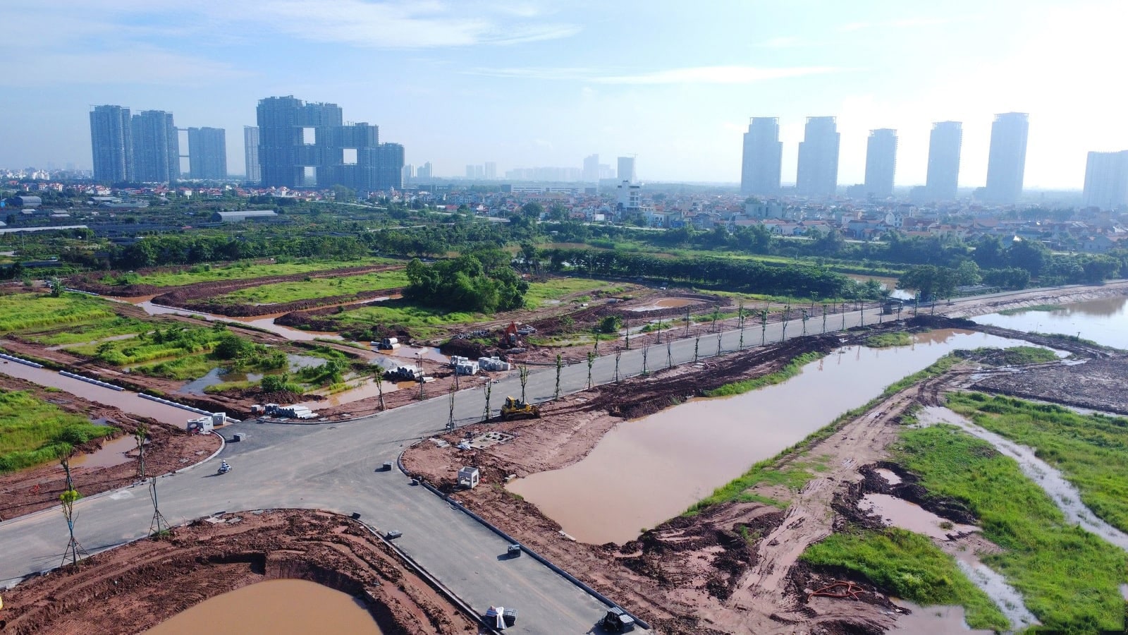 Ảnh chụp từ trên cao công trường dự án Alluvia City vào tháng 7 năm 2025, cho thấy các tuyến đường nội khu đã trải nhựa, một số khu vực đã phân lô, có cây xanh trồng dọc đường và nhiều máy móc thi công đang hoạt động; phía xa là khu đô thị với nhiều tòa nhà cao tầng.